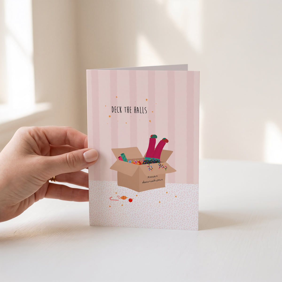 A hand holds a pink striped greeting card featuring an illustration of a girl’s legs with colourful socks sticking out of a box with the text 'DECK THE HALLS' above. The background is a softly lit, warm-toned indoor setting.