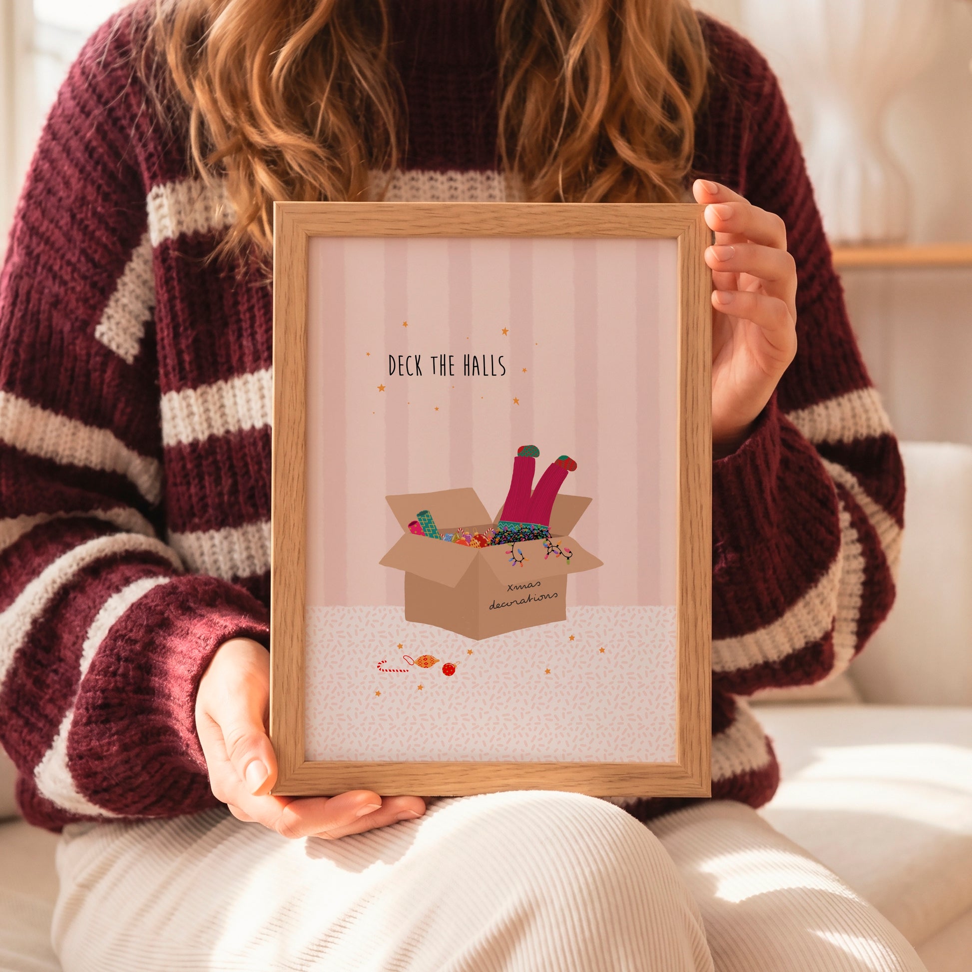A person in a burgundy and cream striped knit sweater holds a light wood framed art print. The print features a pink striped background with the text 'DECK THE BALLS' above an illustration of colorful deck balls spilling out of a cardboard box.