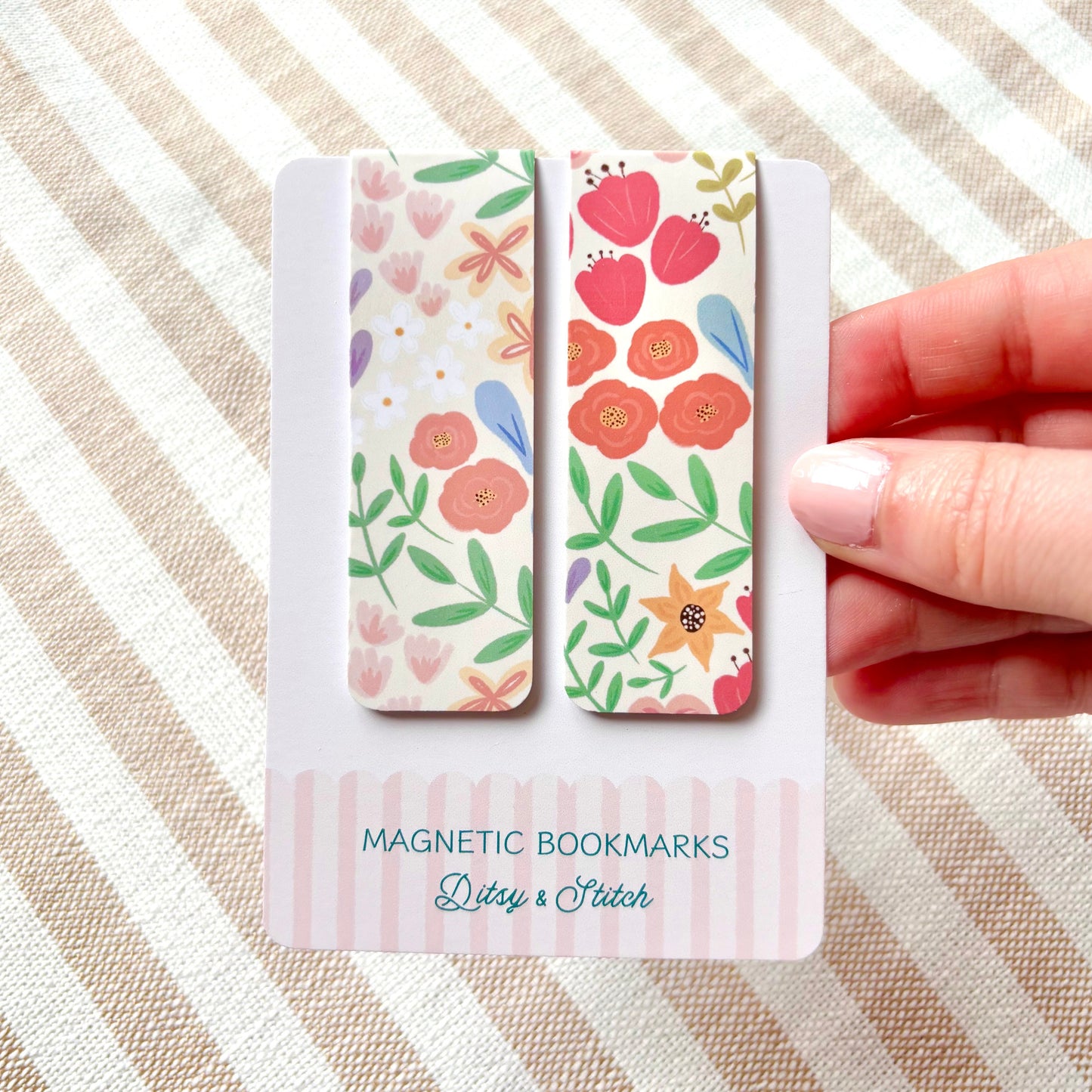 Hand holding two floral magnetic bookmarks with packaging on a striped fabric background