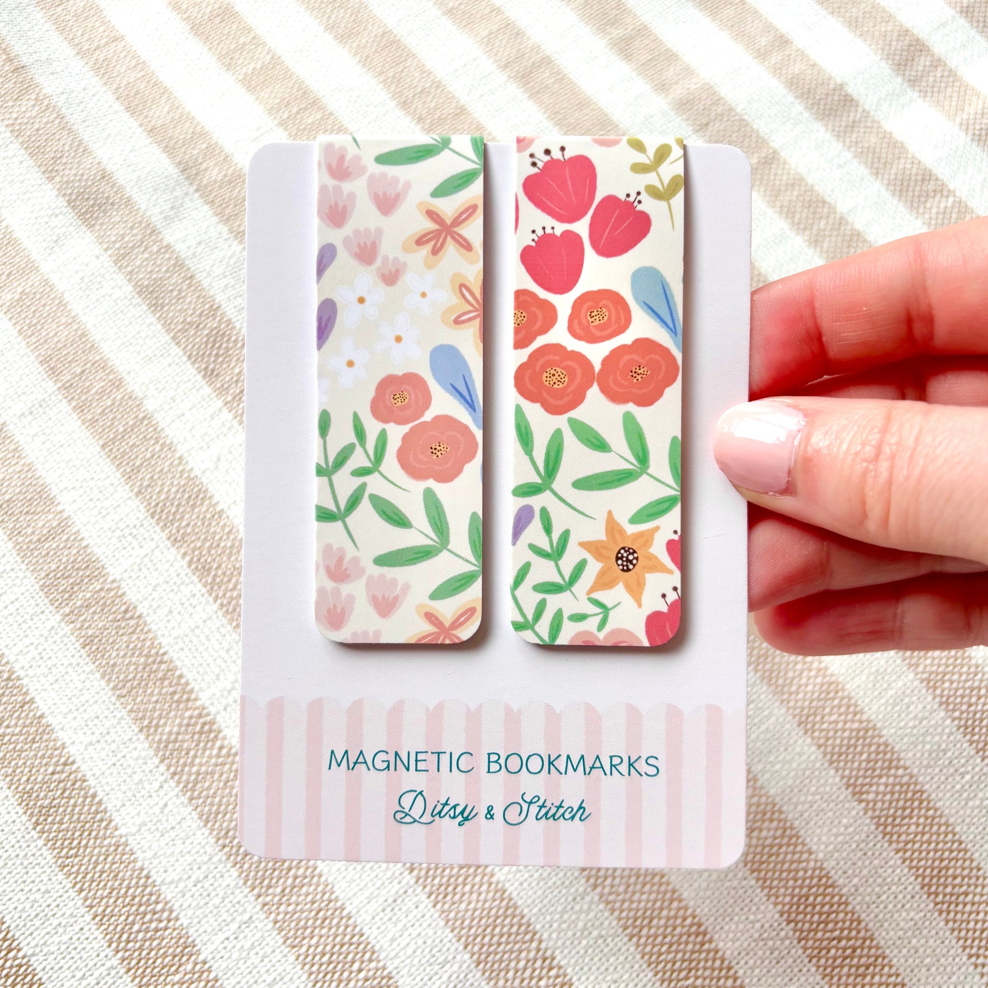 Hand holding two floral magnetic bookmarks with packaging on a striped fabric background