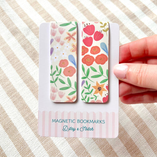 Hand holding two floral magnetic bookmarks with packaging on a striped fabric background