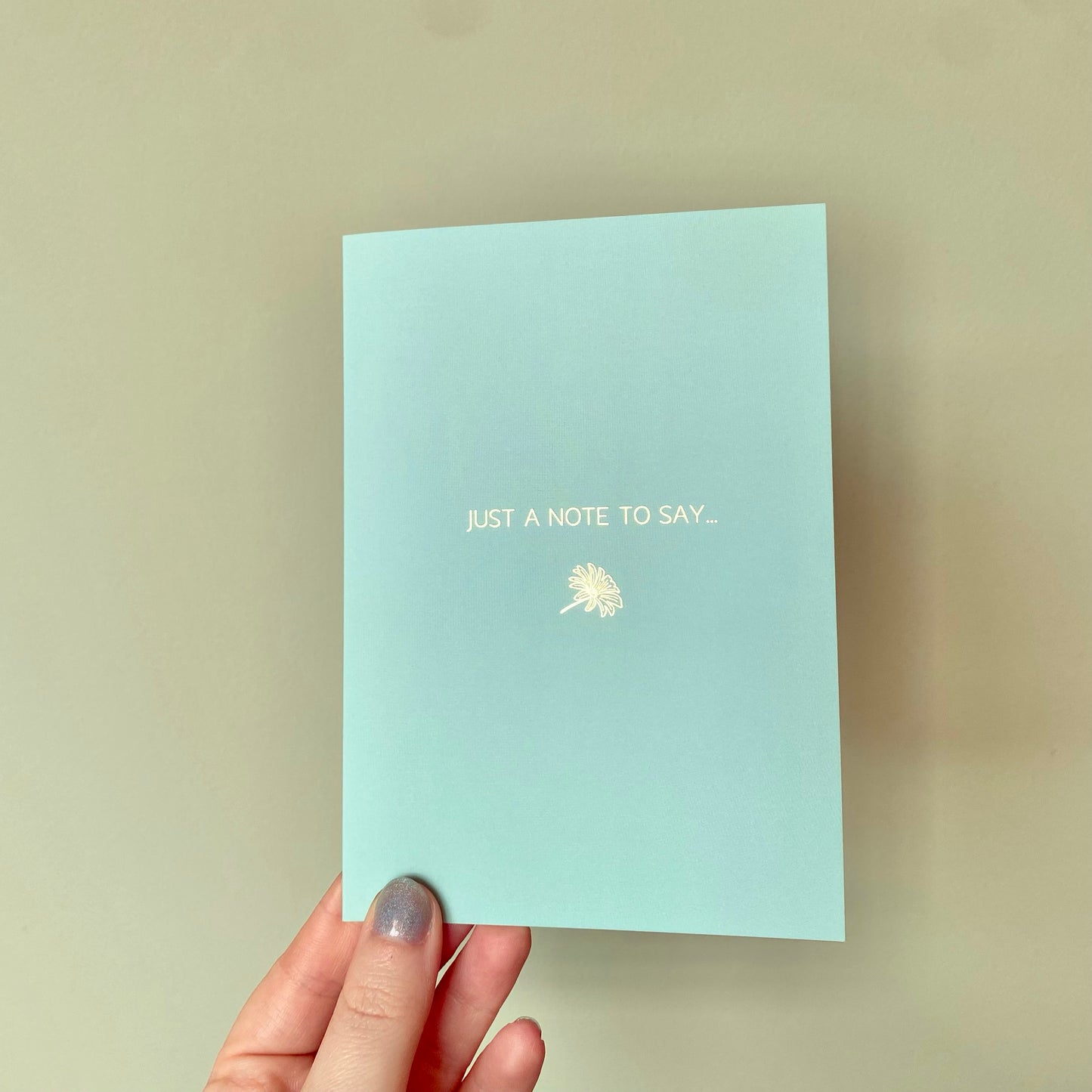 Mint blue card with gold text and daisy design, held by a hand on a green background.