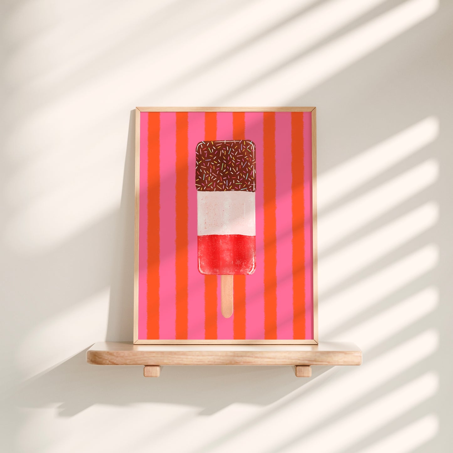 A framed art print featuring a popsicle on a background of pink and orange vertical stripes, displayed on a light wood shelf against a wall with soft window shadow patterns.