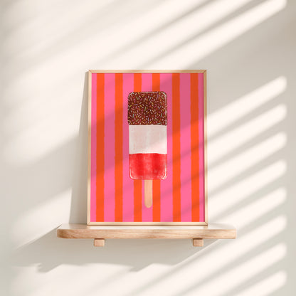 A framed art print featuring a popsicle on a background of pink and orange vertical stripes, displayed on a light wood shelf against a wall with soft window shadow patterns.