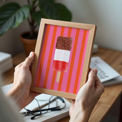 A person holds a light wood framed art print featuring a hand-drawn popsicle on a pink and orange striped background, set in a cosy indoor environment with a desk, plant, and books in soft focus.