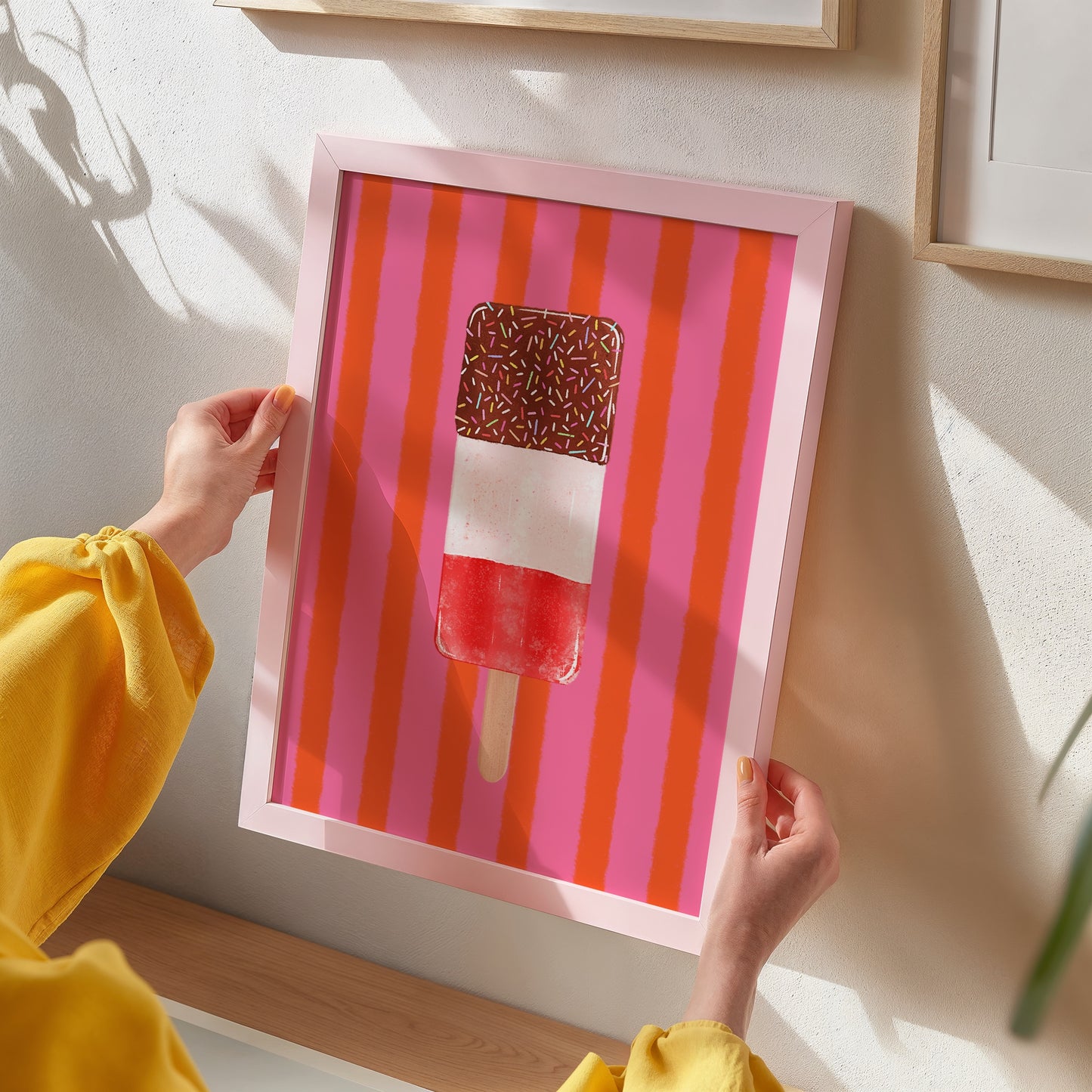A person in a yellow top holds a framed art print featuring a popsicle on a pink and orange striped background, displayed on a light beige wall.