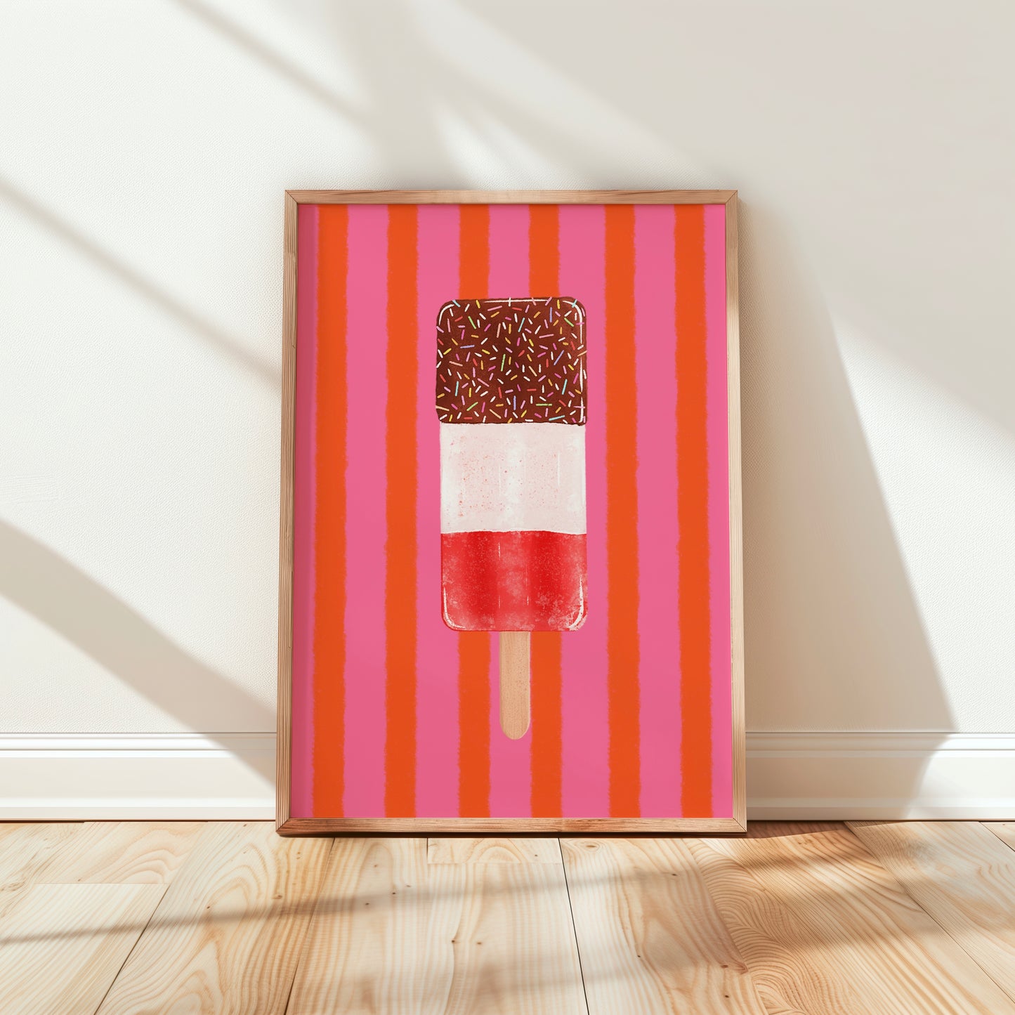 Framed wall art leaning against a white wall on a light wood floor, featuring a stylized popsicle on a pink and orange striped background.