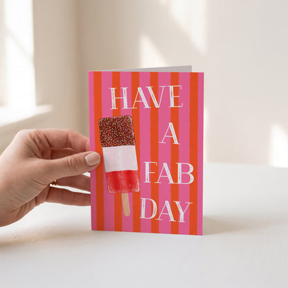 A hand holds a greeting card with a pink and orange striped background, white text reading 'HAVE A FAB DAY', and an ice lolly illustration on the front, set against a softly lit indoor background.