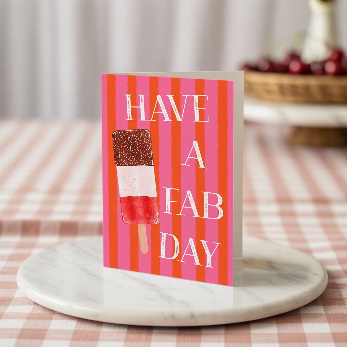 A greeting card with a pink and orange striped background, featuring an ice lolly illustration and the text 'HAVE A FAB DAY' in white. The card is displayed on a round white marble tray over a pink and white checkered tablecloth.
