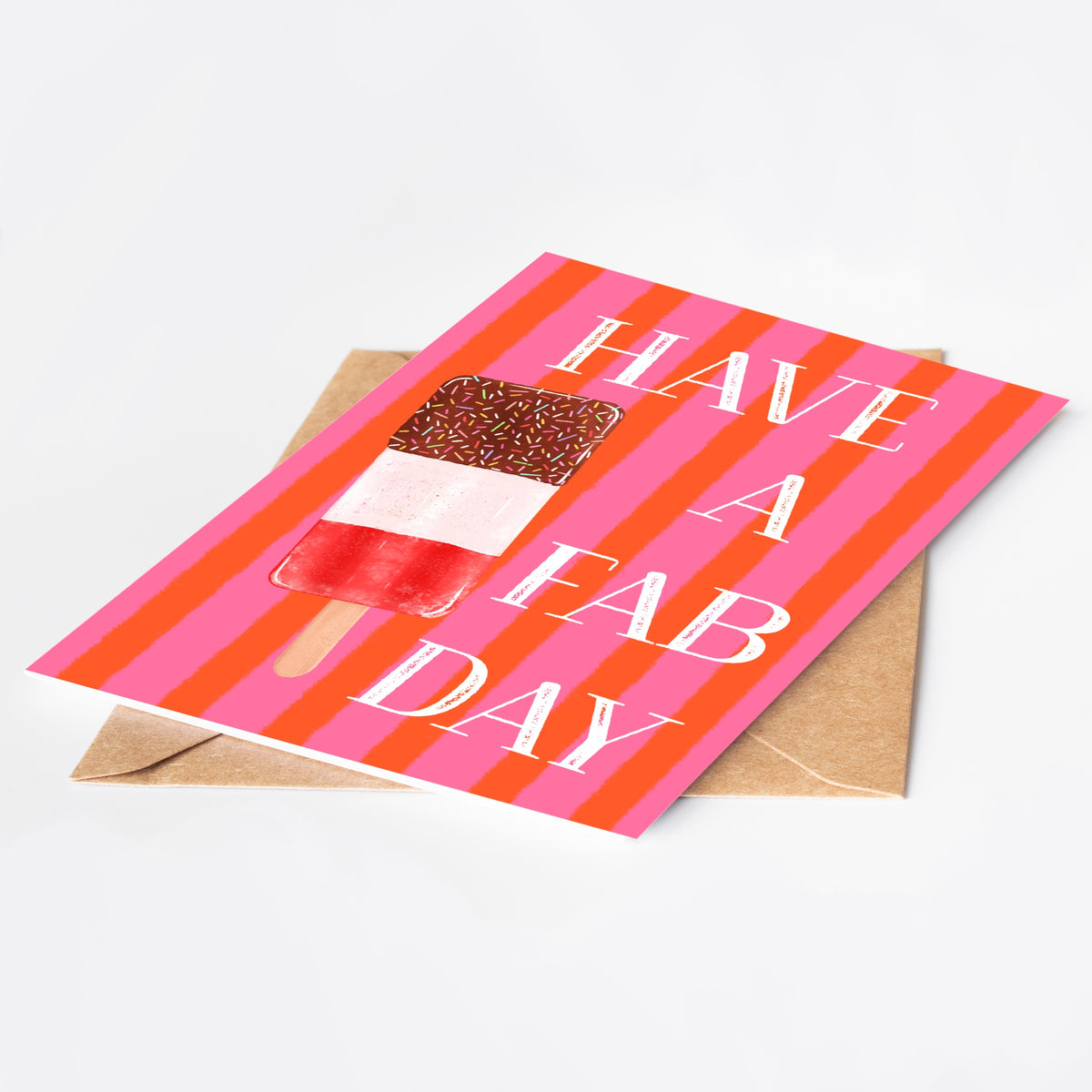 A greeting card with a pink and orange striped background, featuring an ice lolly and the text 'HAVE A FAB DAY' in white. The card is displayed at an angle on top of a brown kraft envelope against a clean white background.