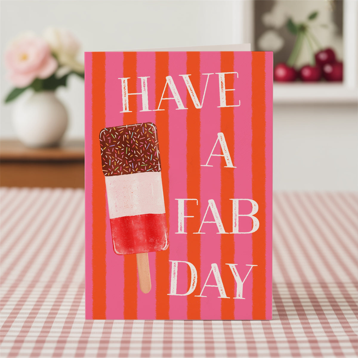 A greeting card standing upright on a red-and-white checkered tablecloth, featuring the phrase 'HAVE A FAB DAY' in white serif text over vertical pink and orange stripes, with an ice lolly in red, white, and brown on the left side.