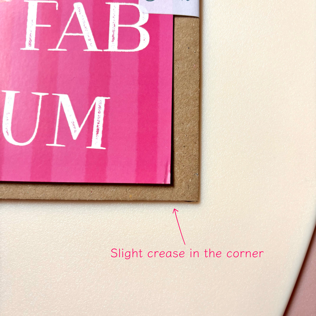 Close-up of a pink printed card with white lettering on a kraft brown backing, placed on a light beige textured surface. Pink text at the bottom reads 'Slight crease in the corner' with an arrow pointing to the card's corner.