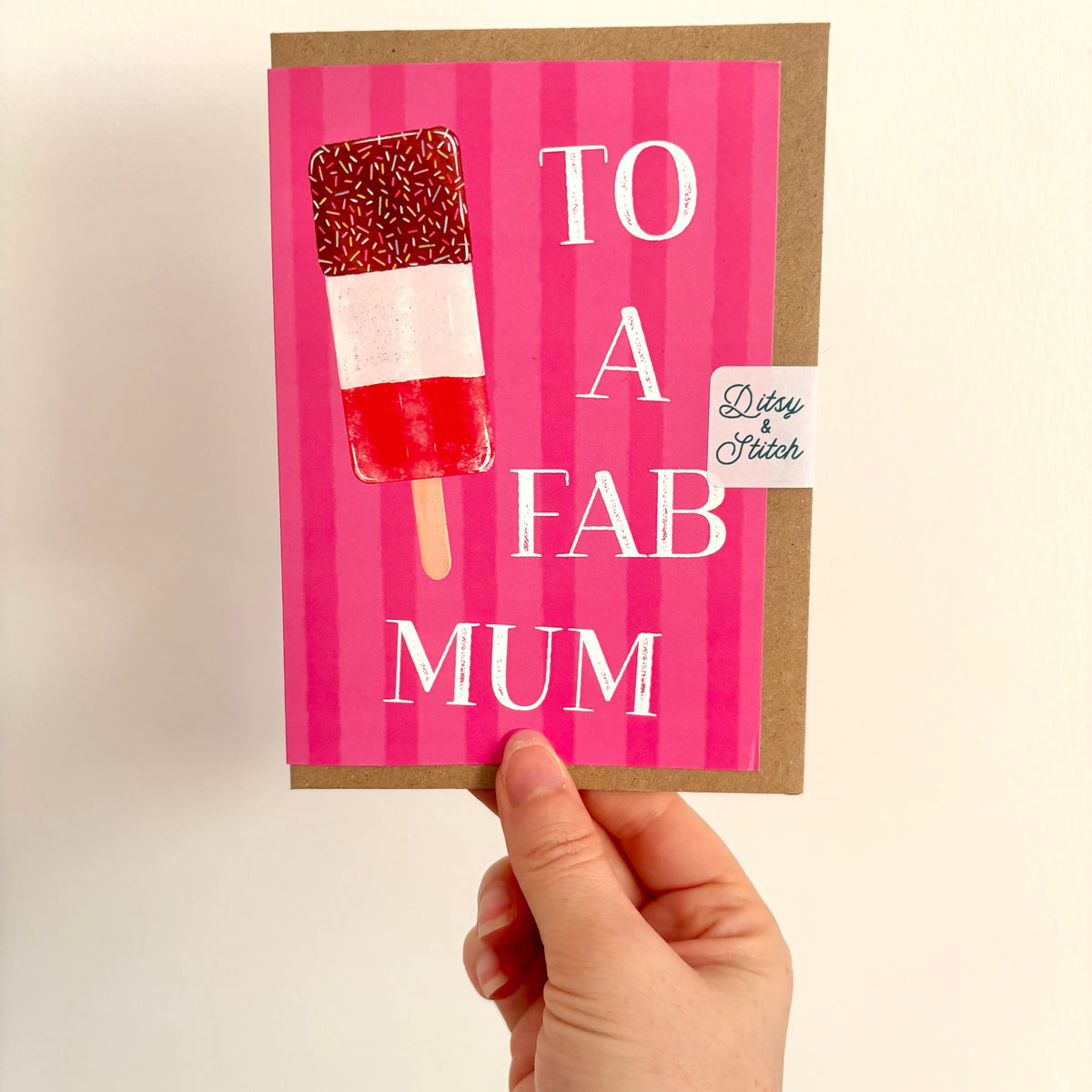 A hand holding a greeting card that says 'TO A FAB MUM' in white text on a bright pink striped background, with an illustration of a red, white, and pink ice lolly. The card has a brown kraft envelope behind it and a small 'Ditsy & Stitch' brand tag.
