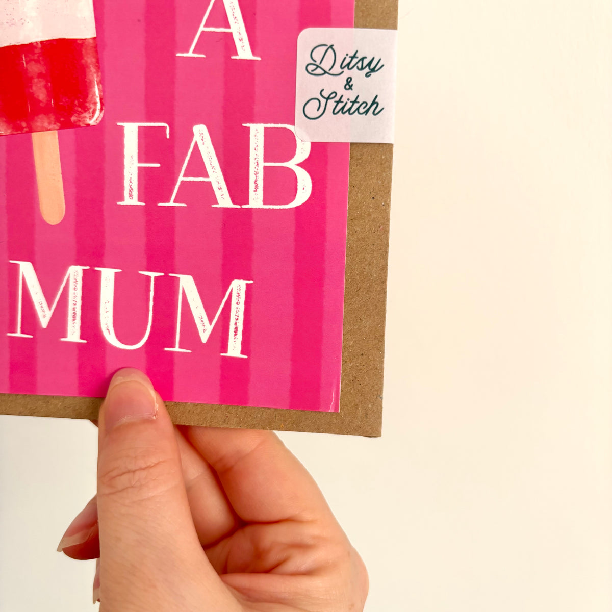 A hand holding a pink greeting card with the words 'A FAB MUM' in white distressed lettering, an ice lolly illustration, and a small 'Ditsy & Stitch' sticker, against a plain cream background.