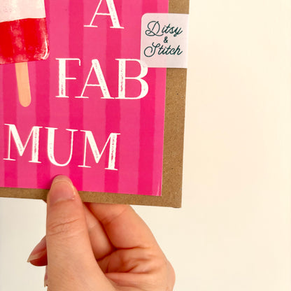 A hand holding a pink greeting card with the words 'A FAB MUM' in white distressed lettering, an ice lolly illustration, and a small 'Ditsy & Stitch' sticker, against a plain cream background.