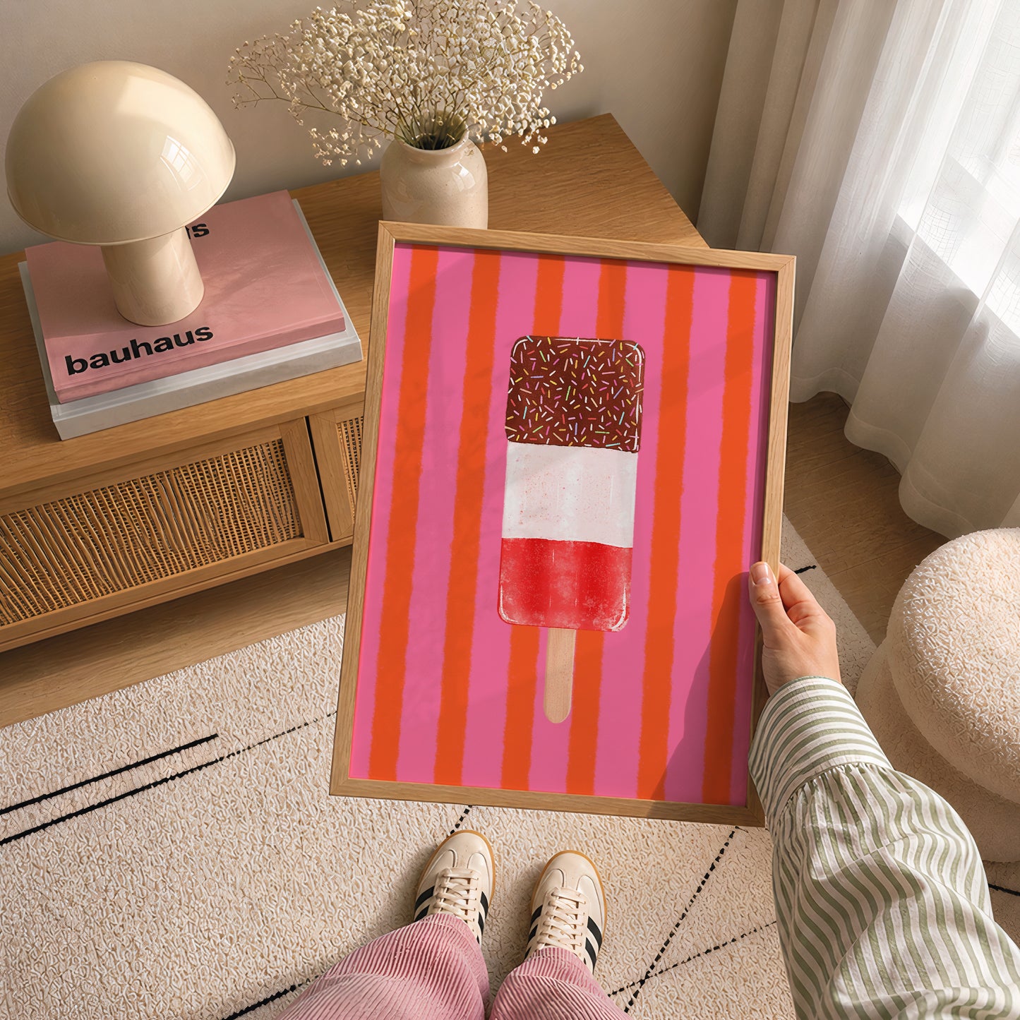 A person holds a framed art print featuring a colourful popsicle on a pink and orange striped background. The scene is set in a cozy, modern room with a wooden side table, a mushroom-shaped lamp, and a patterned rug.