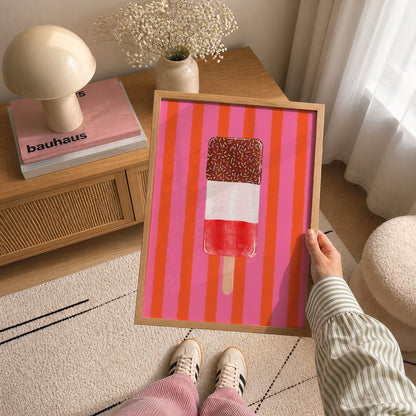 A person holds a framed art print featuring a colourful popsicle on a pink and orange striped background. The scene is set in a cozy, modern room with a wooden side table, a mushroom-shaped lamp, and a patterned rug.