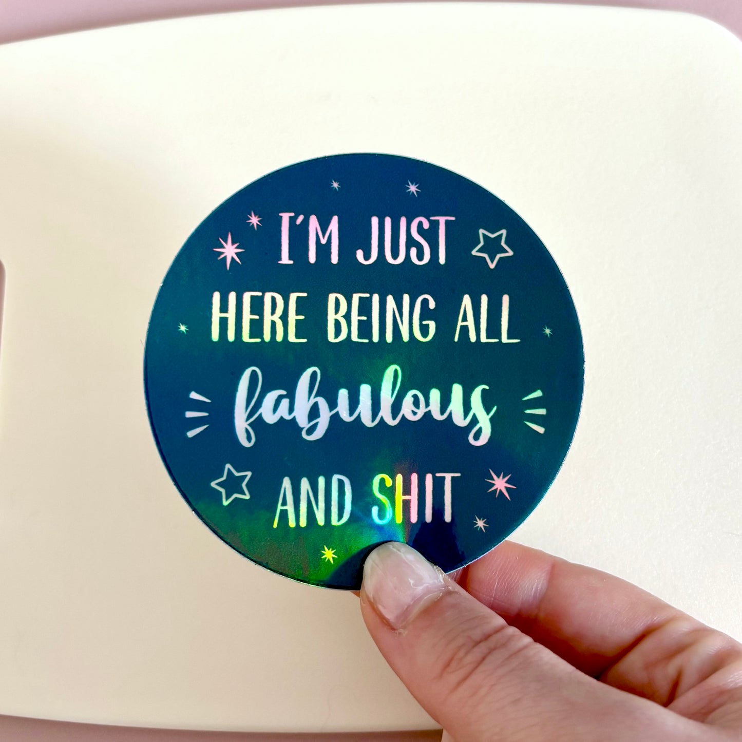 A round holographic sticker held by a hand, featuring the text 'I'M JUST HERE BEING ALL fabulous AND SHIT' in white and iridescent lettering on a dark blue background with star details.