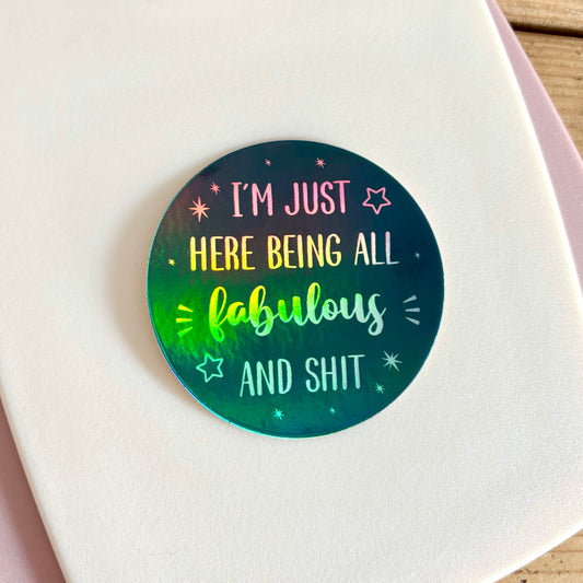 A round sticker with a holographic navy background and the text 'I'M JUST HERE BEING ALL fabulous AND SHIT', shown on a white card.