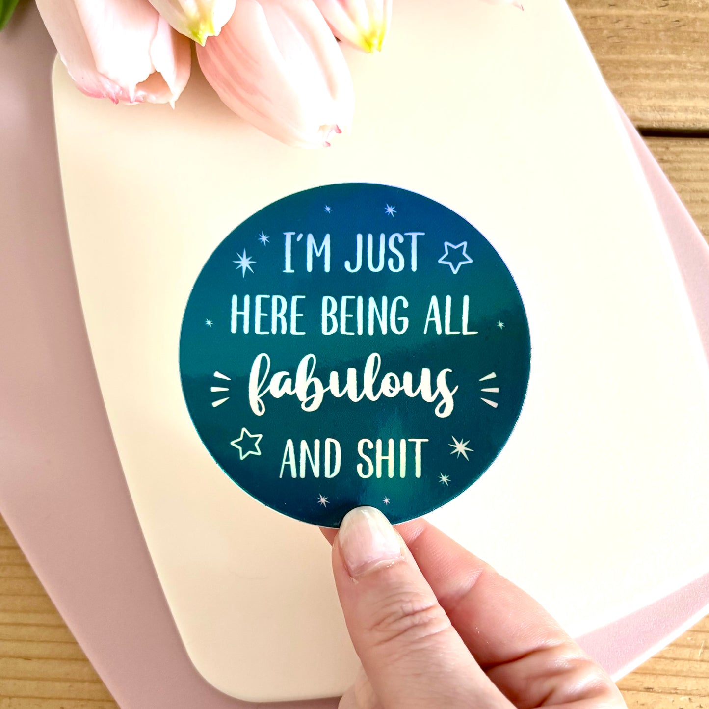 A hand holds a round sticker with the text 'I'M JUST HERE BEING ALL fabulous AND SHIT' in white holographic lettering on a dark teal background. The sticker is shown on a cream-colored card, with pink tulips and a wooden surface in the background.