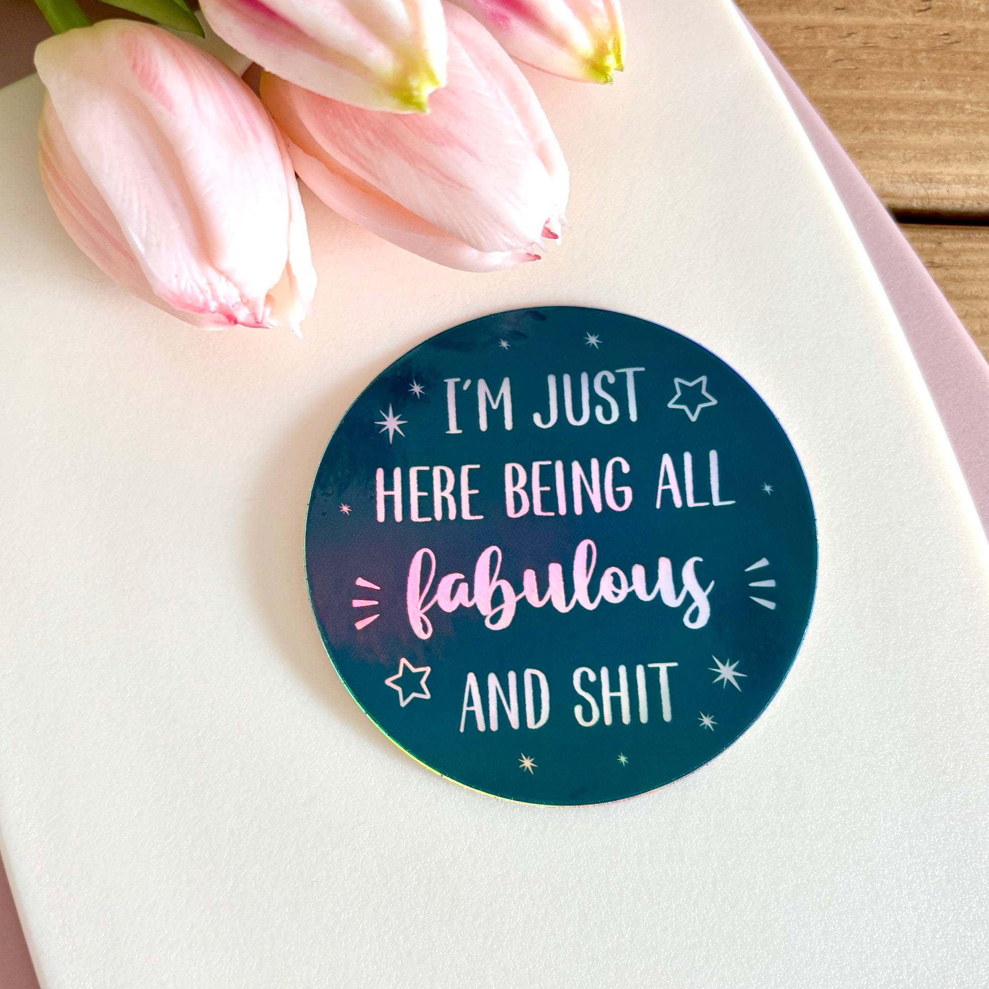 A round sticker with the text 'I'M JUST HERE BEING ALL fabulous AND SHIT' in white holographic lettering on a dark teal background, styled on a white card with pink tulips.