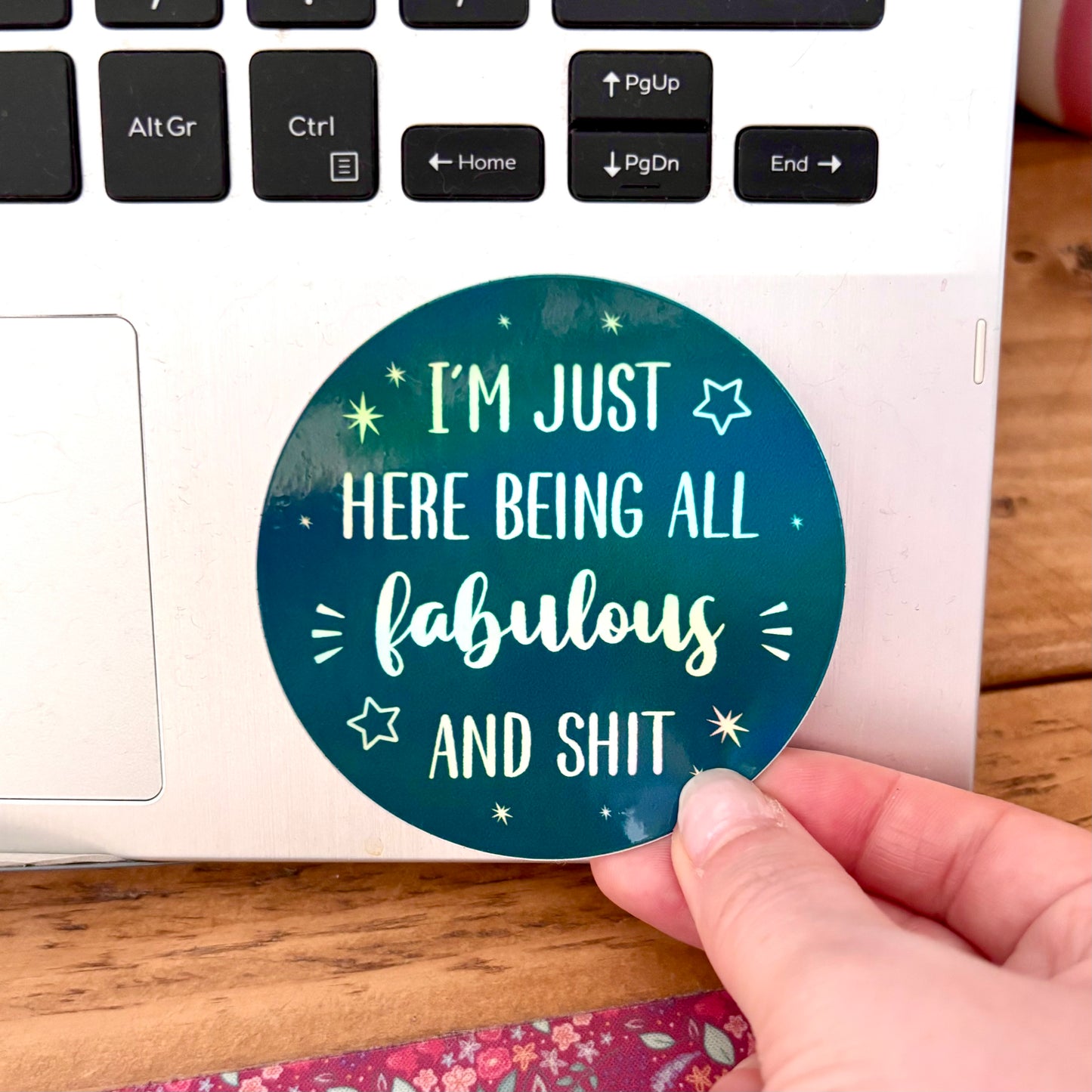 A hand is peeling a round sticker off the corner of a white laptop. The sticker has a dark teal background with white text that reads, 'I'M JUST HERE BEING ALL fabulous AND SHIT' surrounded by small white stars.