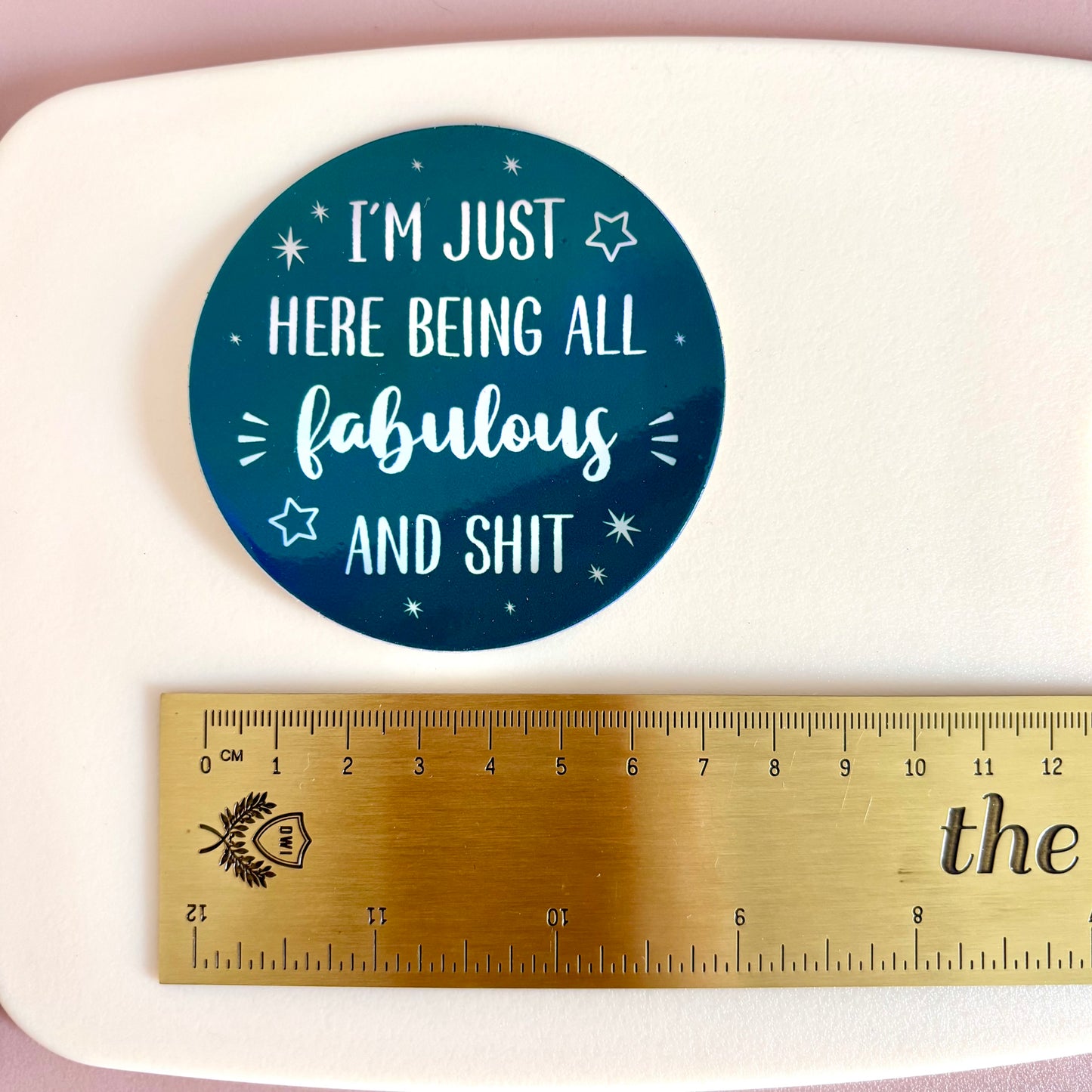 A round teal sticker with white text reading 'I'M JUST HERE BEING ALL fabulous AND SHIT' is shown above a gold metal ruler on a white surface to indicate size.