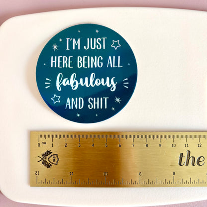 A round teal sticker with white text reading 'I'M JUST HERE BEING ALL fabulous AND SHIT' is shown above a gold metal ruler on a white surface to indicate size.