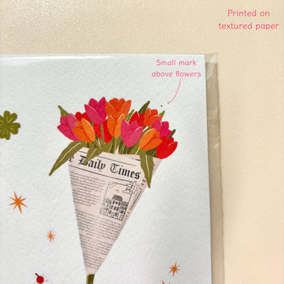A close-up of a print on a warm beige textured background. The design shows a bouquet of tulips wrapped in a newspaper cone, with pink, red, orange, and green flowers, plus small decorative star and flower motifs.