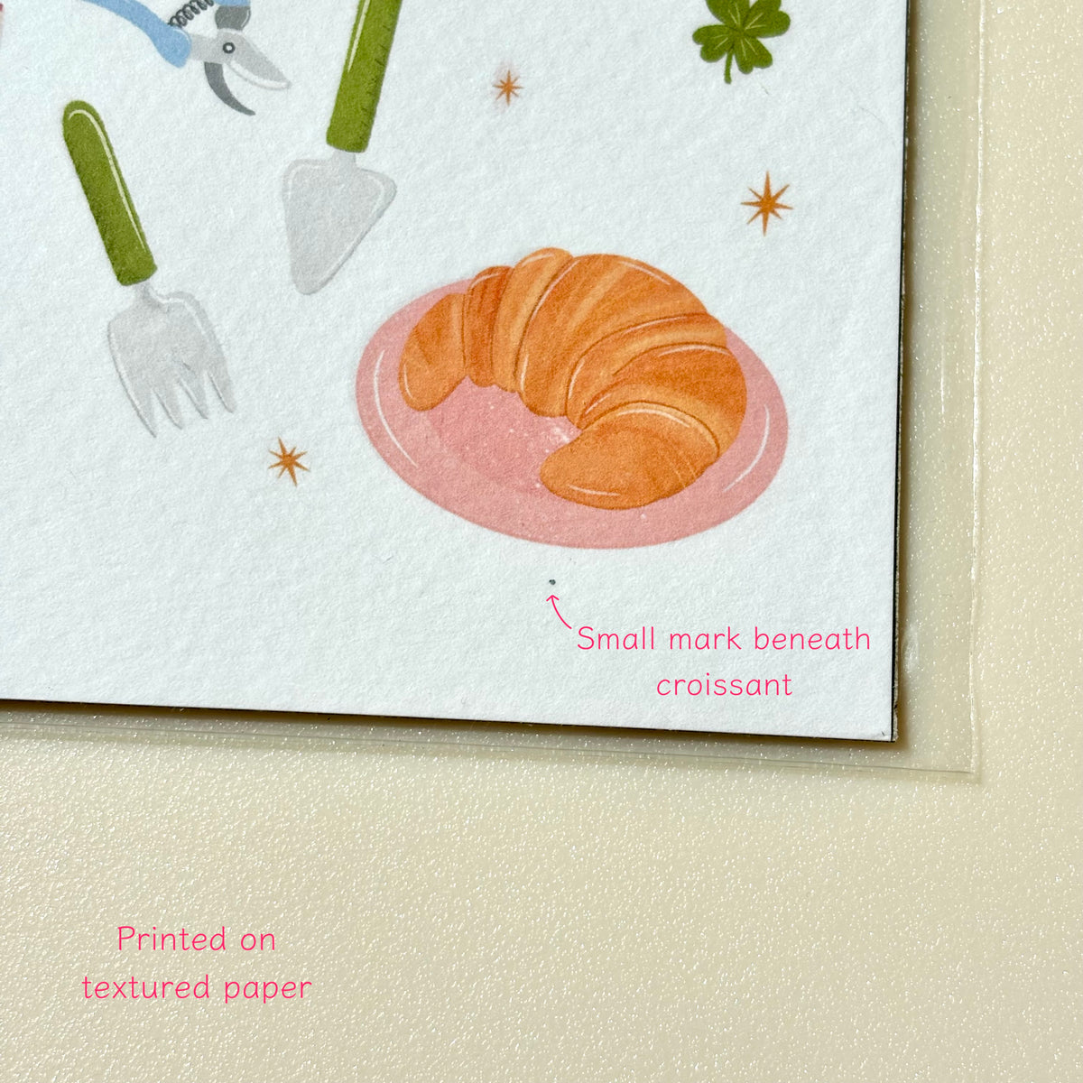 Close-up of a printed illustration on textured paper showing a croissant on a pink plate, with decorative utensils and small star accents. Pink text reads 'Small mark beneath croissant' and 'Printed on textured paper'.