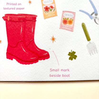 A close-up of a print showing a pair of bright red rubber rain boots on textured white paper, with small garden-themed illustrations around them and text pointing out a small mark beside the boot.