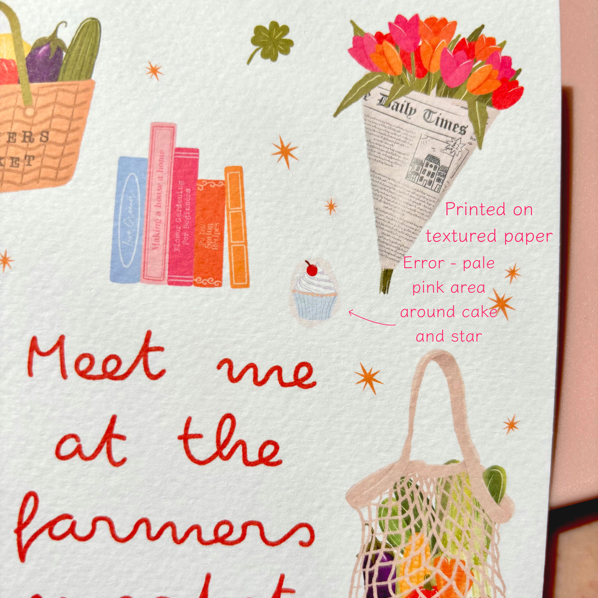 A close-up of a print featuring whimsical illustrations on textured white paper, including red handwritten text reading 'Meet me at the farmers market,' colourful flowers in a newspaper cone, books, a basket of vegetables, a cupcake, and a fruit bag. Pink annotation text points out a printed error on the paper.