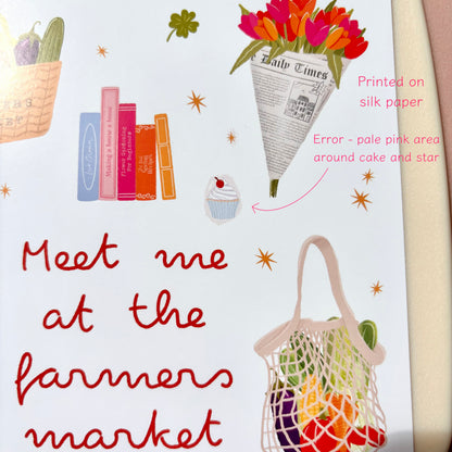 A white art print featuring whimsical illustrations and the phrase 'Meet me at the farmers market' in red script. The design includes a basket of vegetables, colourful books, a bouquet of flowers wrapped in a newspaper cone, a cupcake, and a mesh bag of fruit.