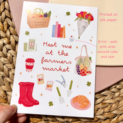 An art print held in a hand, featuring illustrated farmer’s market items and the text 'Meet me at the farmers market' in red script. The print is photographed on a woven straw mat.