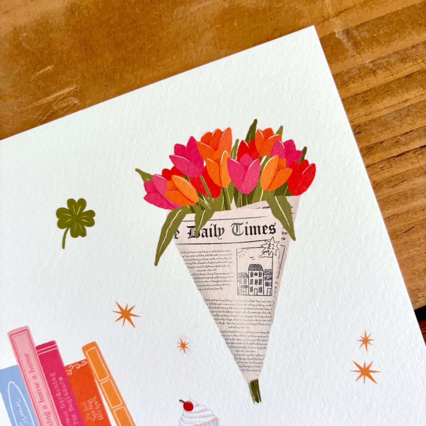 A close-up of a white textured art print featuring a bouquet of tulips wrapped in newspaper, with decorative stars, a clover, books, and a cupcake, placed on a wooden surface.