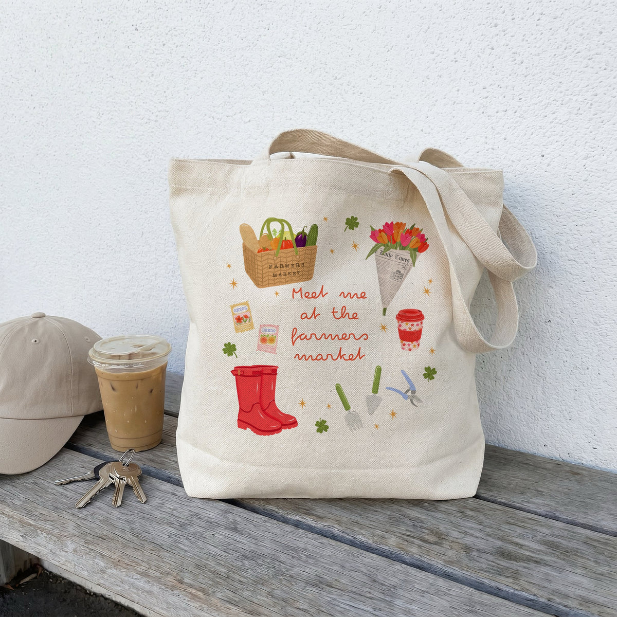 A beige canvas tote bag with a colourful illustrated design sits on a weathered wooden bench against a white wall. The bag features drawings of a basket of vegetables, flowers, red rain boots, gardening tools, and the phrase 'Meet me at the farmers market.