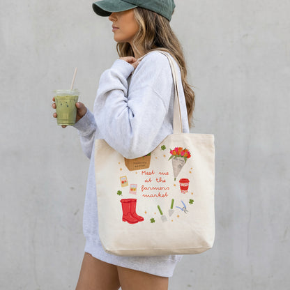 A woman in a light grey sweatshirt and green cap carries a cream canvas tote bag with a colourful market-themed print that reads 'Meet me at the farmer's market.' She is holding an iced green drink against a light grey wall.