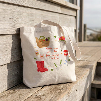 A cream canvas tote bag with a hand-painted 'farmers market' design, leaning against a rustic wooden boardwalk railing.