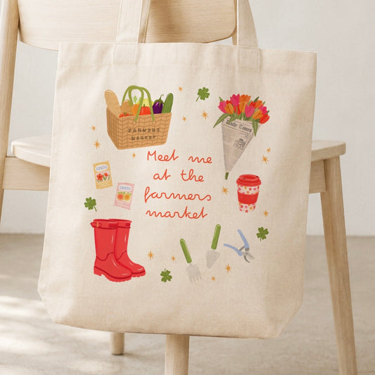 A cream canvas tote bag with a colourful 'Meet me at the farmers market' illustrated print, hanging from a light wood chair in a bright, neutral room.