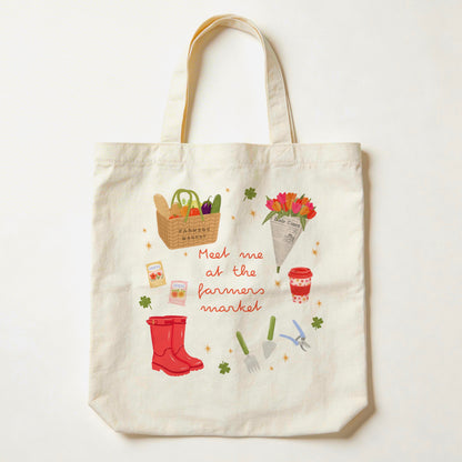 An off-white canvas tote bag with a colourful 'Meet me at the farmers market' illustration, featuring red rain boots, gardening tools, flowers, and a basket of vegetables, shown against a plain white background.