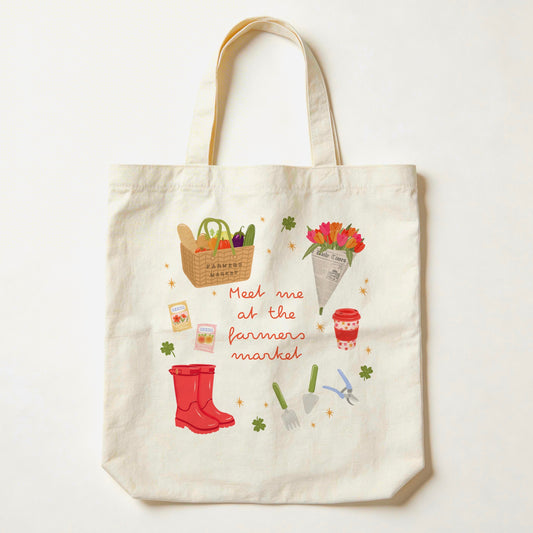 An off-white canvas tote bag with a colourful 'Meet me at the farmers market' illustration, featuring red rain boots, gardening tools, flowers, and a basket of vegetables, shown against a plain white background.