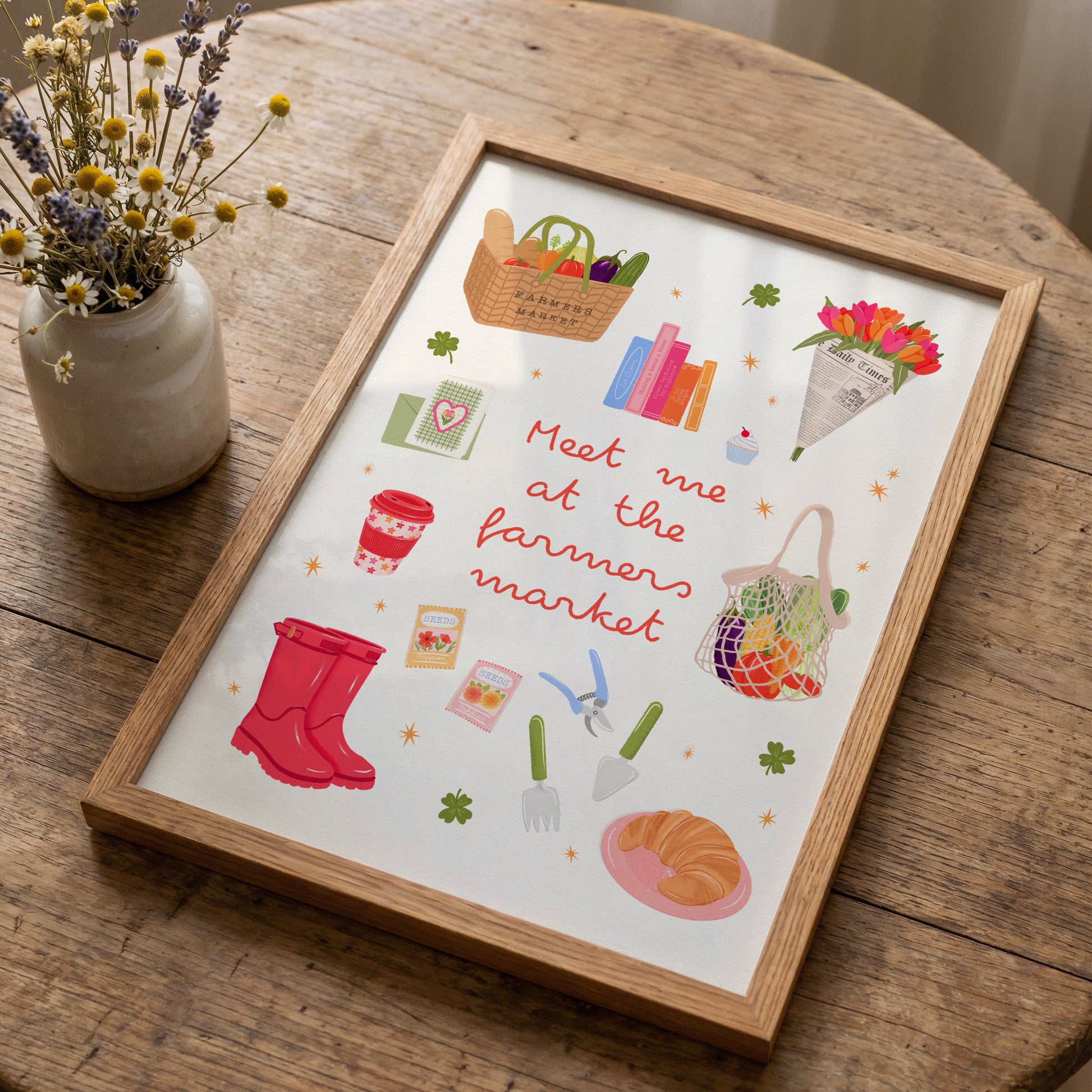 A framed art print featuring a whimsical 'Meet me at the farmers market' illustration, styled on a rustic wooden table next to a small vase of dried flowers.