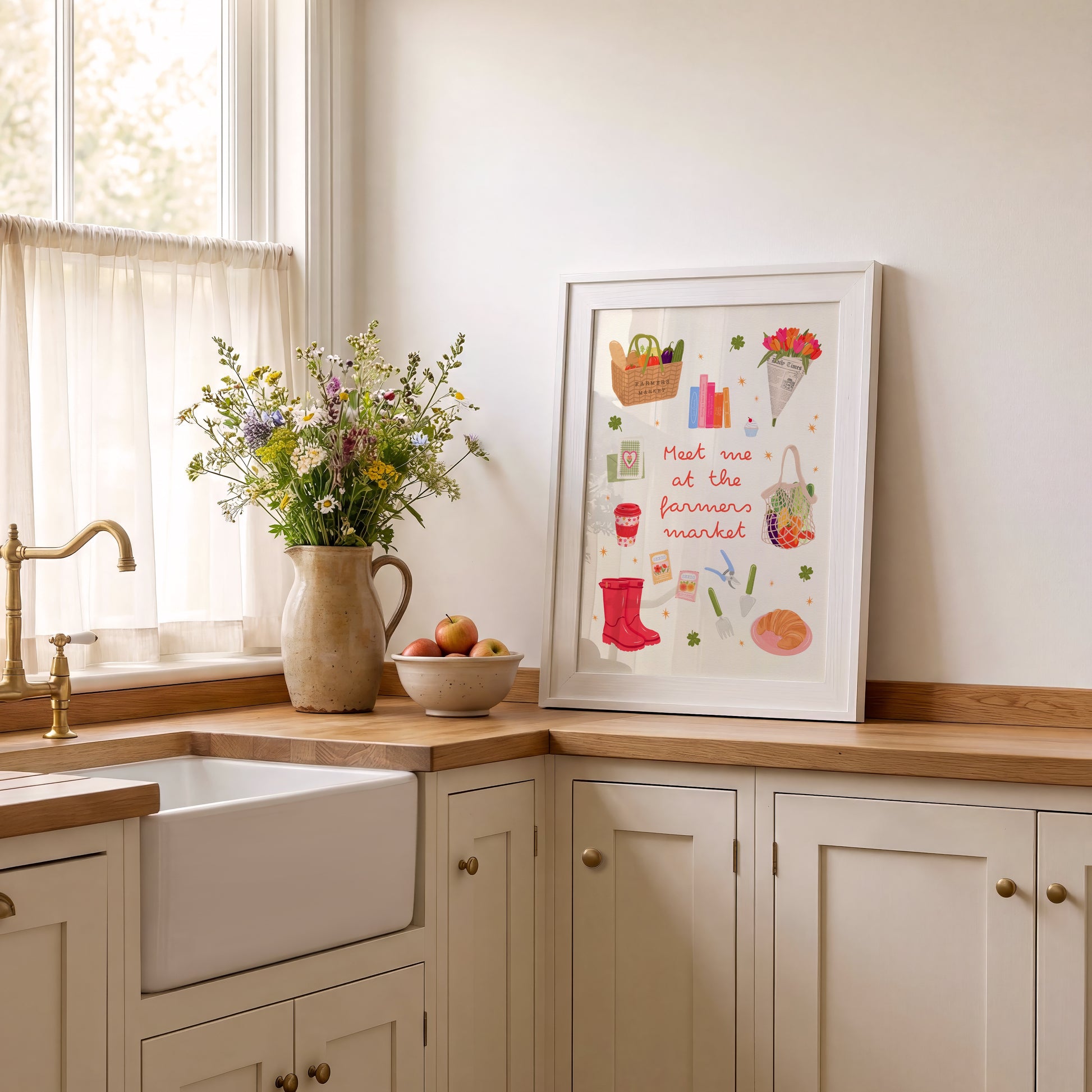 A framed art print featuring a whimsical market illustration and the text 'Meet me at the farmer's market' leaning on a light wood kitchen counter next to a farmhouse sink and a vase of flowers.
