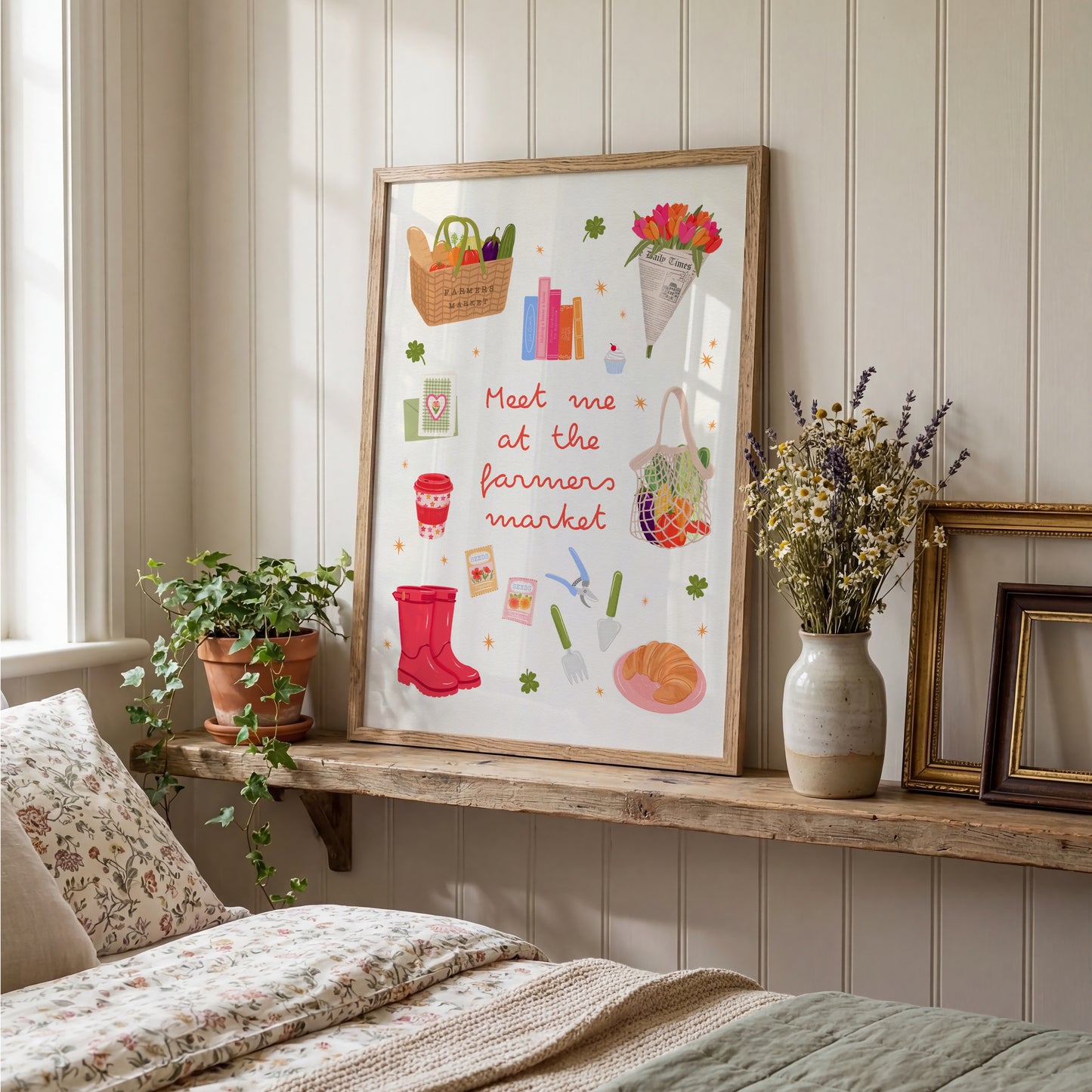A framed art print featuring a whimsical 'Meet me at the farmers market' illustration with red rain boots, flowers, and market items, styled on a wooden shelf in a cozy bedroom setting.