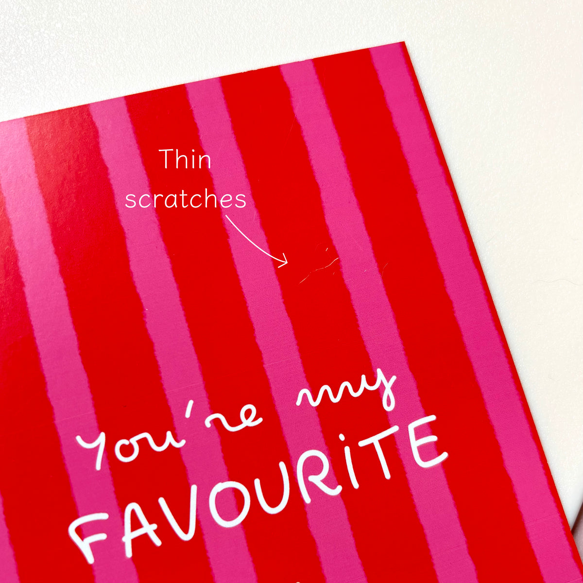 A close-up of a red greeting card with pink vertical stripes and the handwritten text 'You're my FAVOURITE' in white. A small scratch is pointed out with an arrow and the words 'Thin scratches' on the card surface.