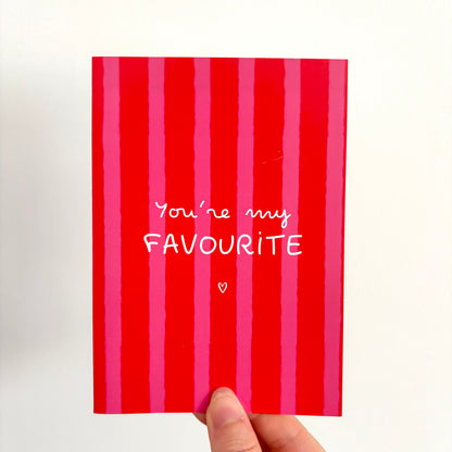A hand holding a red and pink vertically striped greeting card that reads 'You're my FAVOURITE' in white text, against a plain white background.