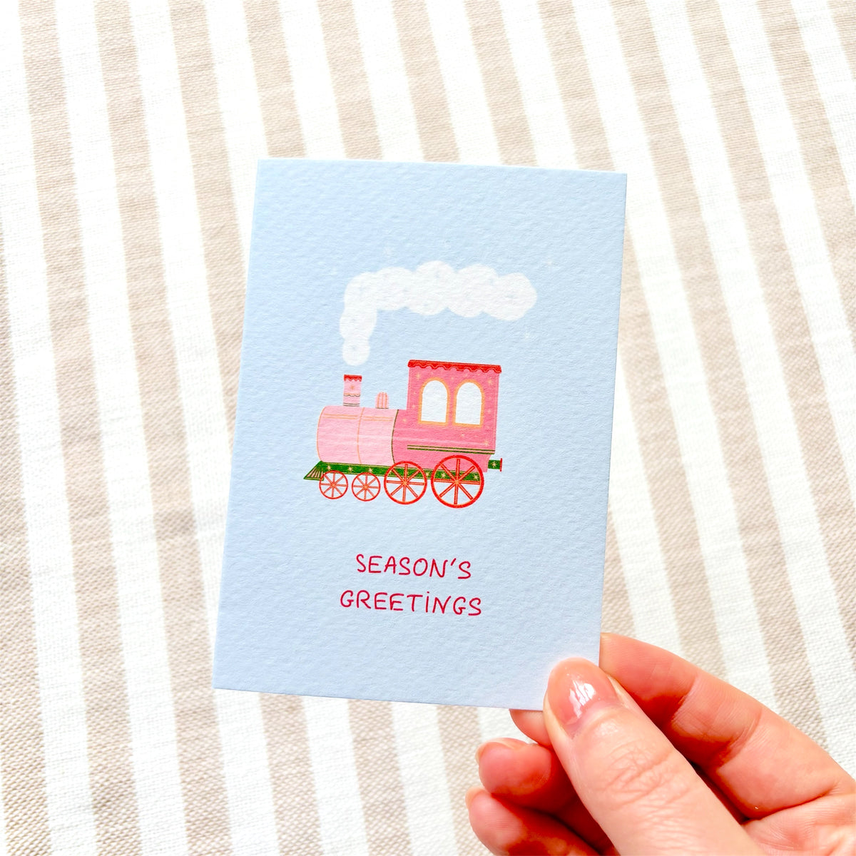 A hand holding a light blue greeting card with a pink steam train illustration and the words 'SEASON'S GREETINGS' on a beige and white striped fabric background.