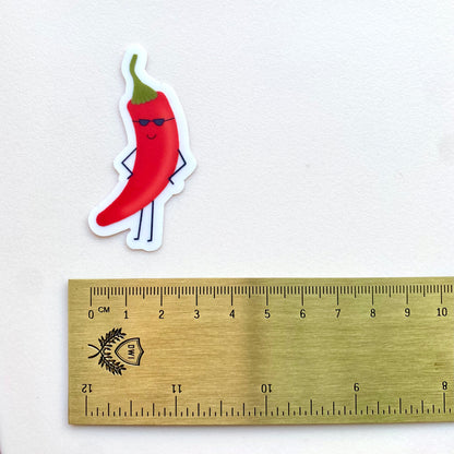 Transparent vinyl sticker with a cartoon red chili pepper with sunglasses shown with a gold ruler for scale against a white background