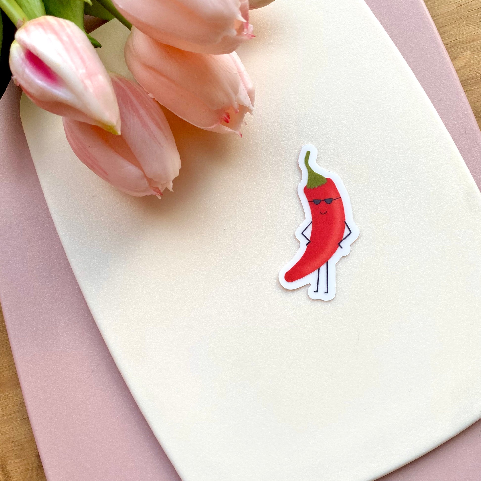 Transparent vinyl sticker with a cartoon red chili pepper with sunglasses shown with pink tulips for scale