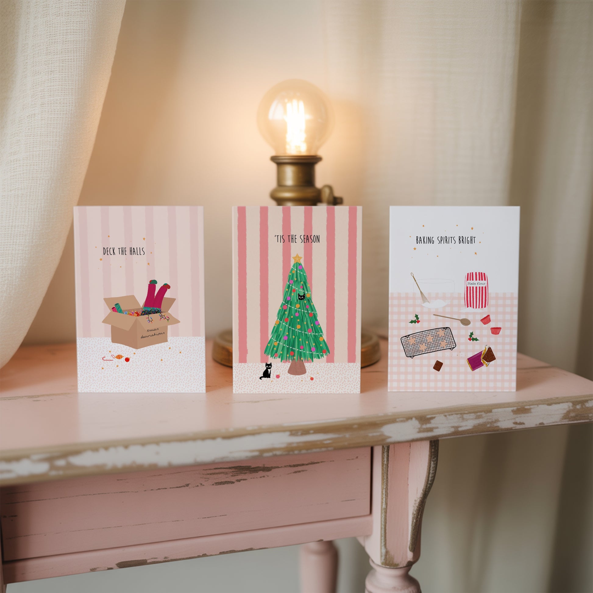 Three pink Christmas-themed greeting cards on a wooden table with a lamp in the background.